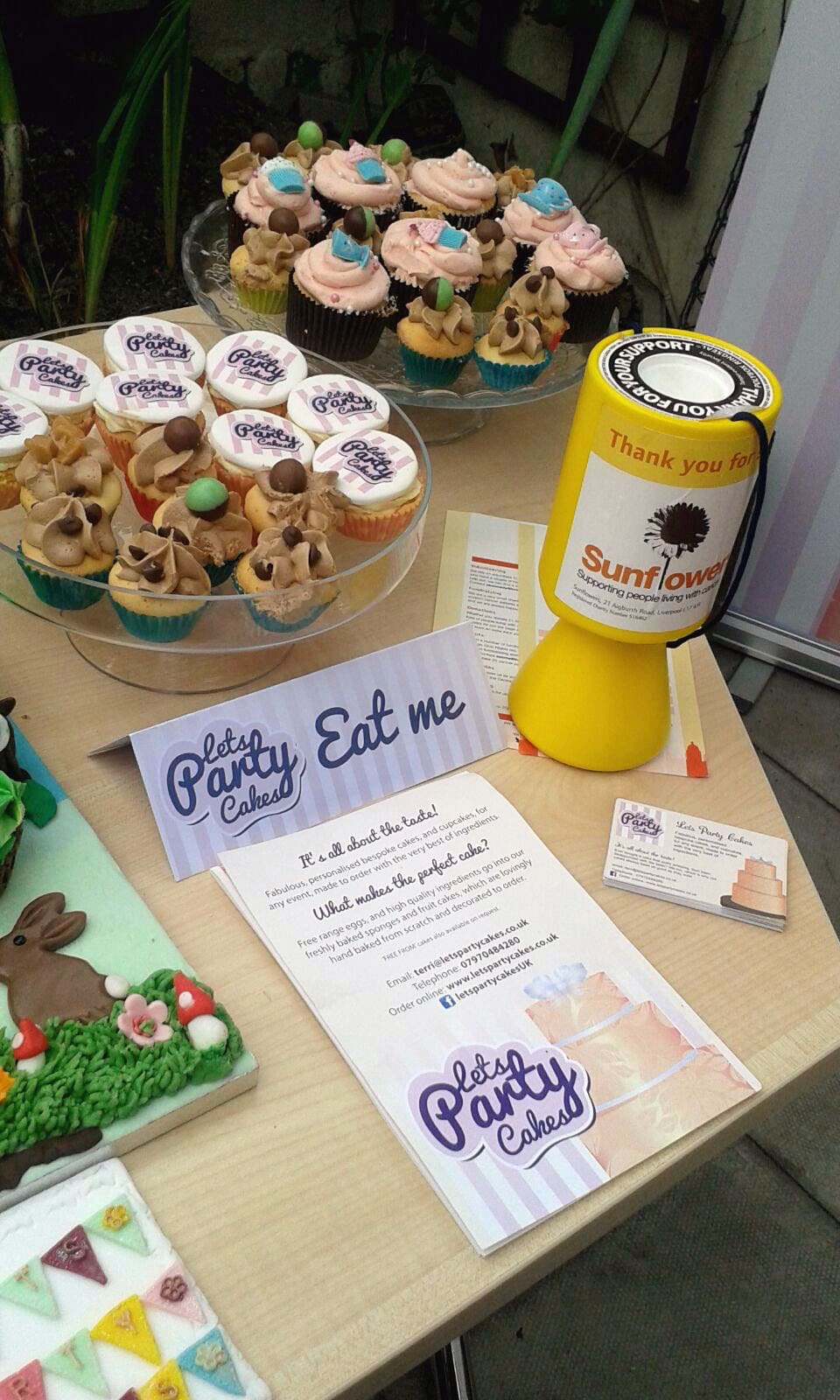Photograph of cake stall at an event with a 'lets party cakes' leaflet and stand that says 'eat me'